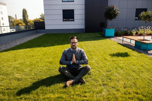 A calm businessman meditating on the meadow in front of the business center