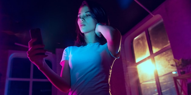 Calm. Cinematic portrait of handsome stylish woman in neon lighted interior. Toned like cinema effects in purple-blue. Caucasian female model using smartphone in colorful lights indoors. Flyer.