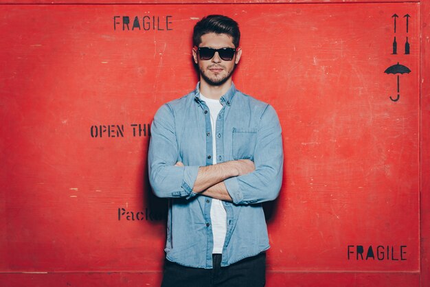 Calm and confident. Handsome young man in sunglasses keeping arms crossed and looking at camera while standing against red background