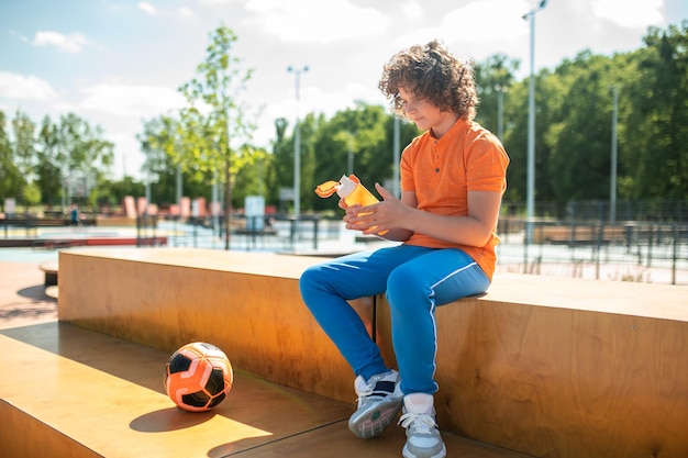 Calm curly-haired boy sitting on the plywood bench in the park and holding an open water bottle in his hands