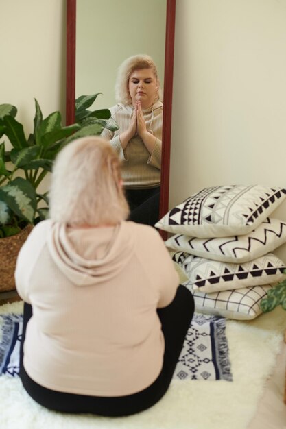 Calm curvy young woman meditating in front of mirror keeping hands in mudra gesture and eyes closed