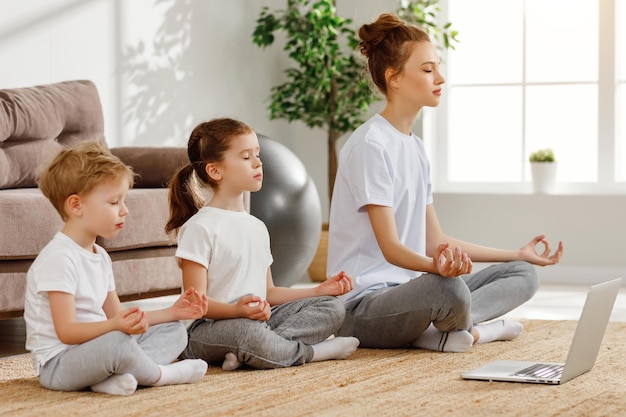 Calm freelance mother with little daughter and song in similar\
outfits sitting all together in lotus pose and meditating in living\
room