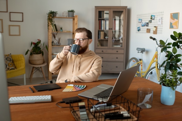Спокойный красивый бородатый дизайнер сидит за столом с компьютерами и пьет кофе в домашнем офисе