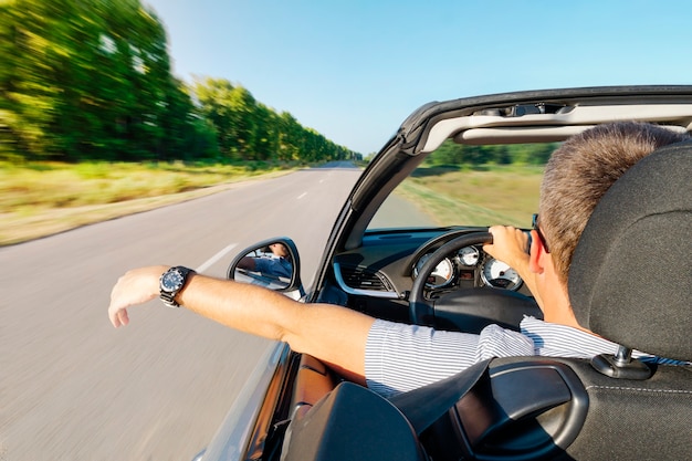穏やかな幸せな成功した男が車の手に座って道路で車を運転しています。高価なコンバーチブルの中の若い男は、美しい夏の自然を背景に、街の外の道路を運転しています。