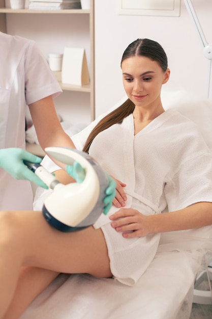 Calm lady feeling glad and smiling while improving the condition of her skin and having special device on the leg