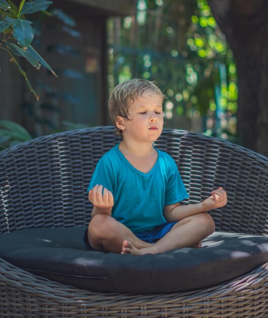 Calm little male boy sit on couch outside garden practice yoga Young generation modern child rest relieve negative emotions with closed eyes in lotus pose at sunrise Mindfulness meditation concept