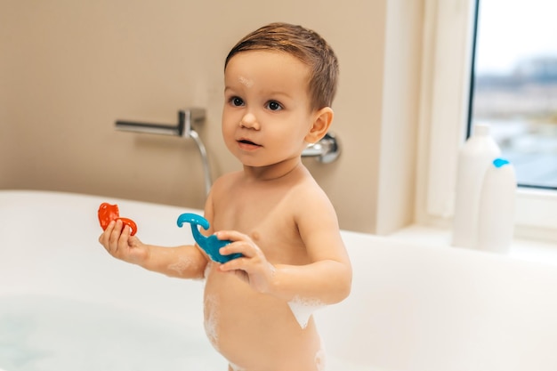 Calm little male child with rubber toys in his hands standing in the bath full of foam