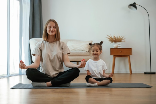 Calm in lotus pose Young woman with little girl are doing yoga at home