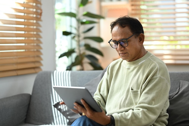 Calm mature man wearing eyeglasses reading online news on\
digital tablet while resting on couch in bright living room