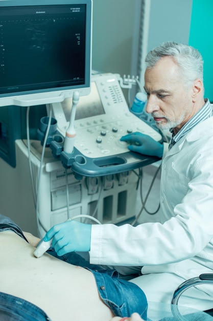 Calm medical worker in a white coat sitting in front of an ultrasound apparatus and touching the belly of a patient with a transducer