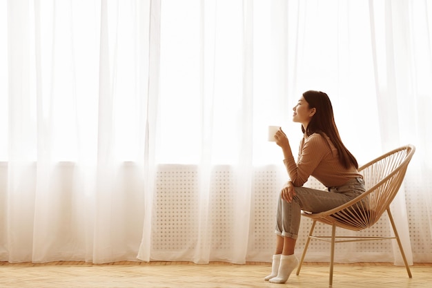 冷静な朝の女の子が現代の椅子に座って窓の横の景色の前でコーヒーを楽しんでいます