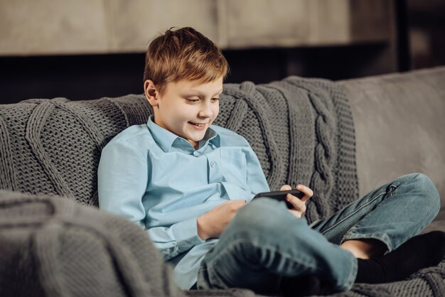 Calm pastime. Upbeat pre-teen boy sitting cross-legged on the sofa and playing on the phone while smiling pleasantly