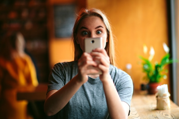 Calm peaceful beautiful young woman take picture using front smartphone camera in cafe or restaurant. Hold device with both hands. Alone in room