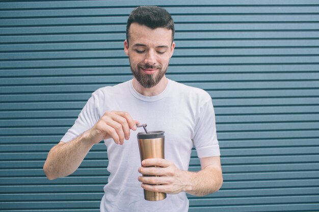 Calm and peaceful man is standing and looking down to thermos with hot drink. He is opening it. Isolated on striped 