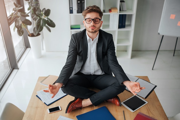 Calm peaceful young man meditating on table in meeting room