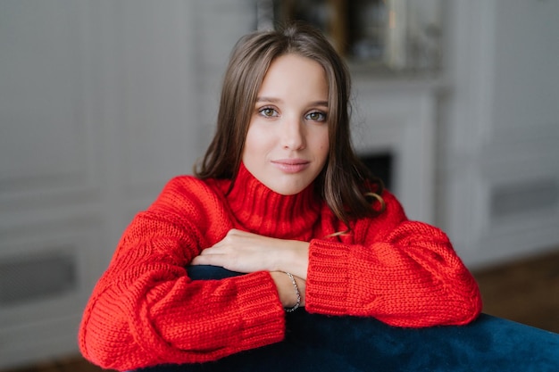 Calm pretty young Caucasian female dressed in warm knitted pullover leans on armchair looks with dark eyes directly at camera poses against domestic interior indoor People and beauty concept