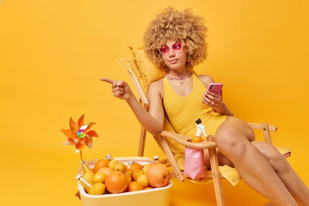 Calm relaxed woman with curly hair dressed in bathingsuit holds\
modern cellphone points away into distance spends summer vacation\
at beach uses portable fridge isolated over yellow background