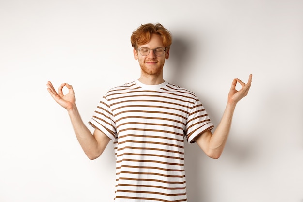 Calm and relaxed young man with red messy hair, spread hands sideways in mudra gesture and smiling, practice yoga or meditating, white background.