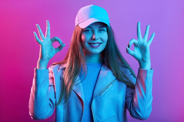 Calm smiling young woman posing indoor, shows okay sign with both hands, wears casual clothing, standing isolated over pink space