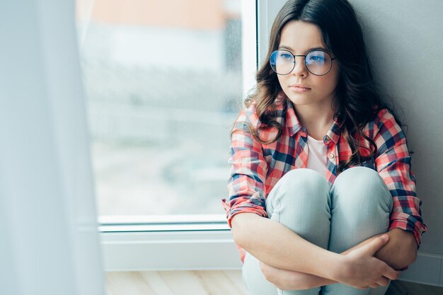 Calm teenage girl wearing glasses embracing her knees while sitting on the floor near the window