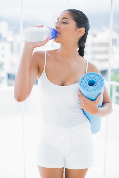 Calm toned brunette holding sports bottle and exercise mat