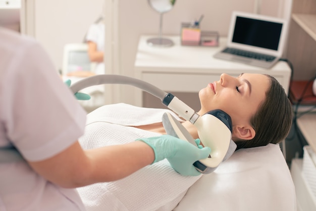 Calm woman lying with her eyes closed and using non-invasive body modeling procedure for improving the condition of her facial skin