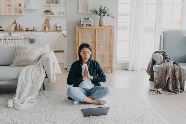 Calm woman practices yoga at laptop has break after working sitting on floor at home Stress relief