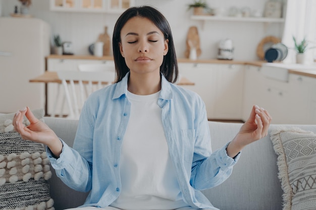 Calm woman practices yoga meditating folded fingers in mudra hand gesture sitting on sofa at home