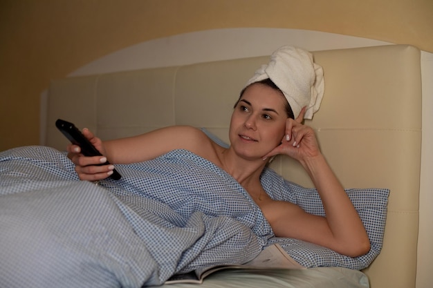 Calm woman with towel on hair lying in bed reading magazine and watching tv using remote control in