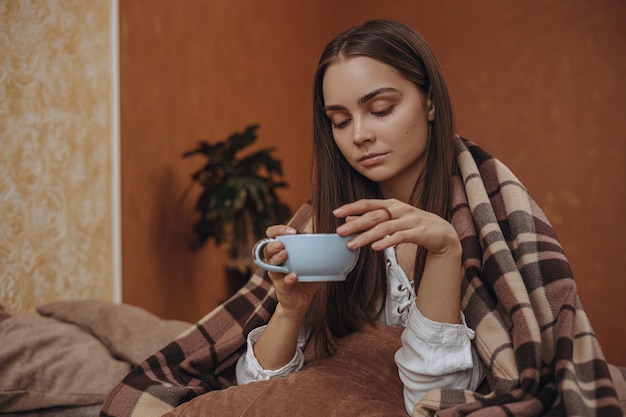 Спокойная женщина, завернутая в теплый плед, пьет горячий чай и отдыхает в уютной комнате дома