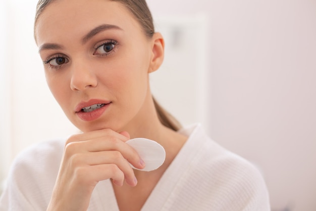 Calm young beautiful lady thoughtfully looking into the distance while holding a cotton pad near her face