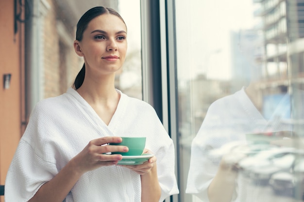 Calm young beautiful lady in a white bathrobe thoughtfully looking into the distance and smiling while standing next to the window with a cup of tea in her hands