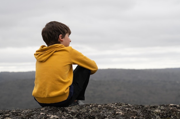 Calm Young boy sitting in the top of a mountain looking at the horizon with copy space