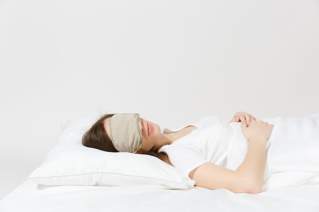 Calm young brunette woman lying in bed with sleep mask on white sheet, pillow, blanket on white wall