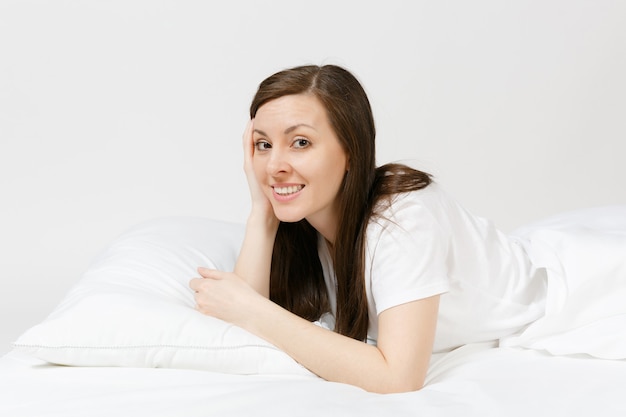 Calm young brunette woman lying in bed with white sheet, pillow, blanket on white wall