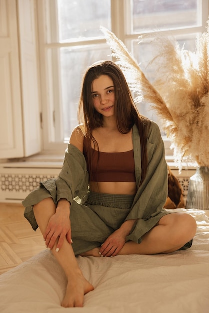 Calm young caucasian woman looks at camera sits crosslegged on floor near window Brunette with long hair wears top shirt and shorts Lifestyle female beauty concept