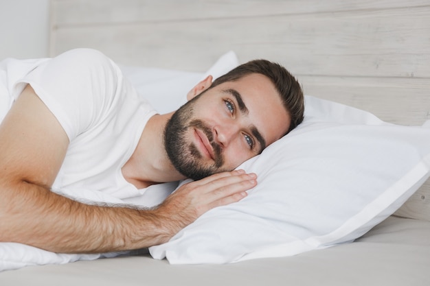 Calm young handsome bearded man lying in bed with white sheet pillow blanket in bedroom at home