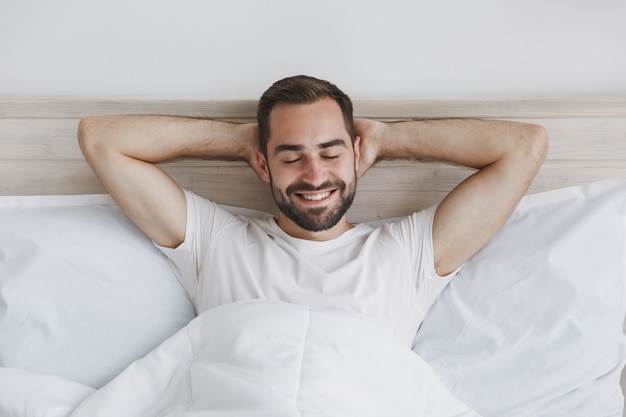 Calm young handsome bearded man lying in bed with white sheet pillow blanket in bedroom at home