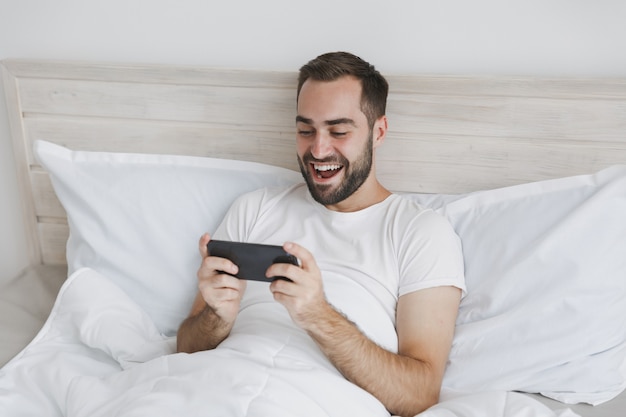 Calm young handsome bearded man lying in bed with white sheet pillow blanket in bedroom at home