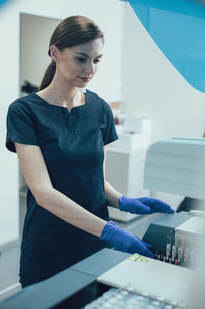 Calm young lady in medical uniform wearing rubber gloves putting test tubes into a modern immunochemistry analyzer