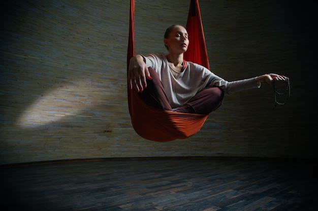 Calm young lady in orange hammock meditating with her eyes closed and holding a string of beads