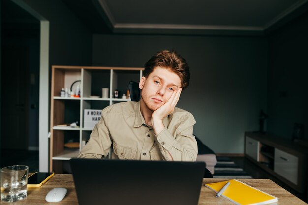 Calm young man in a shirt sitting with a laptop at home at the\
table looking at the camera with a serious face portrait of upset\
freelancer working at home online quarantine concept