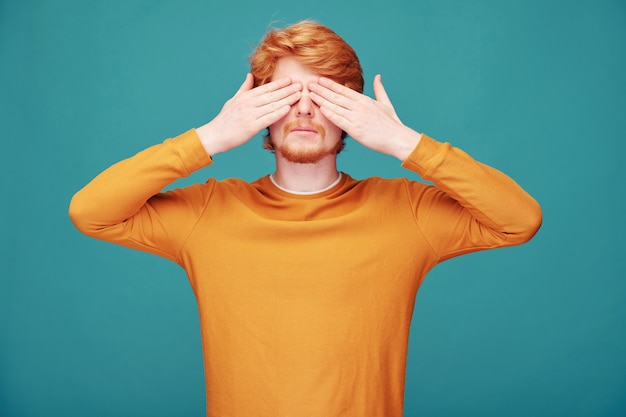 Photo calm young redhead man with beard closing eyes with hands in anticipation of surprise