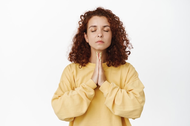 Calm young redhead woman, mindful girl meditating, showing namaste buddha gesture with realxed resting face, yoga pose, standing on white