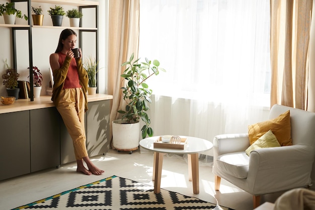 Calm young woman in casual outfit leaning on counter and drinking tea in living room