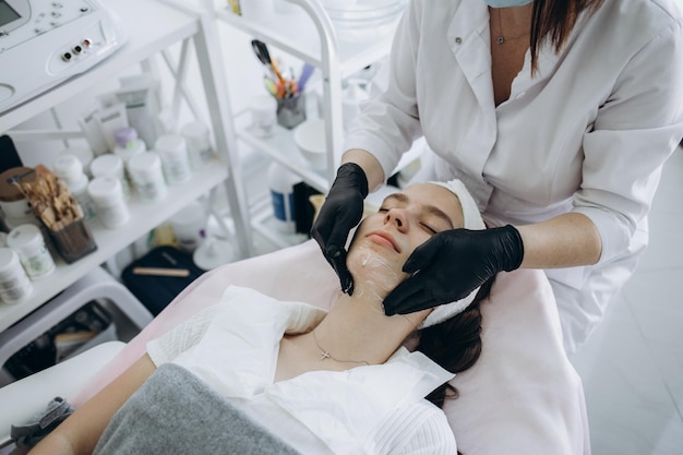 Calm young woman is getting antiaging rejuvenation procedure at beauty salon Beautician hands holding tool under her face Top view