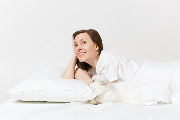 Calm young woman lying in bed with white cute Persian silver chinchilla cat, sheet, pillow, blanket on white wall