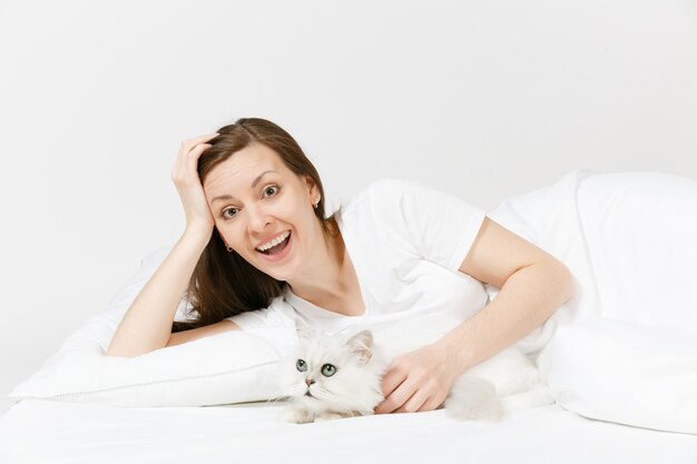 Calm young woman lying in bed with white cute Persian silver chinchilla cat, sheet, pillow, blanket on white wall