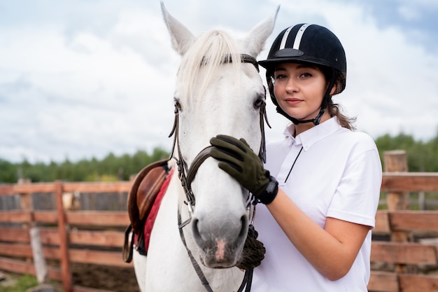 Calm young woman in white polo shirt and equestrian outfit embracing purebred horse while training in rural environment