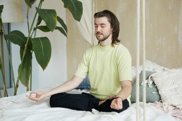 Calmed man sitting on the bed with eyes closed, male in lotus asana and yoga mudra practicing meditation on the bed